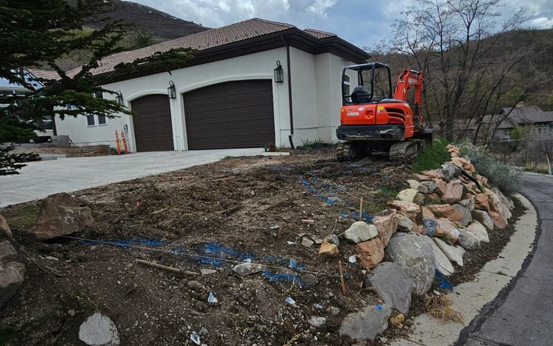 Mini excavator on a cleared hillside lot with boulder edging